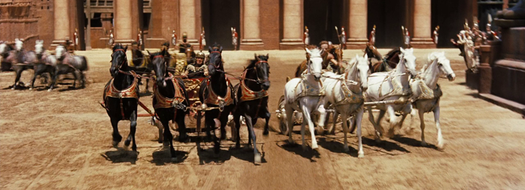 The chariot race in Ben-Hur: a team of four black horses and a team of four white horses pull chariots side by side towards the camera across a sandy arena lined with tall columns