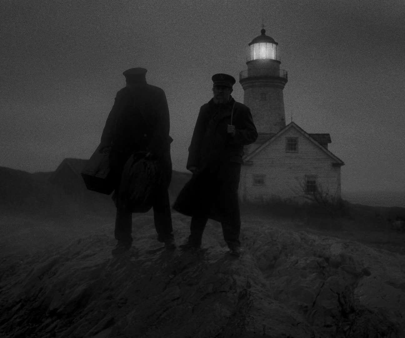 A black-and-white shot of two lighthouse keepers in long coats and caps standing on rocks in the fog, carrying luggage, with the lit lighthouse tower and a white cottage behind them.