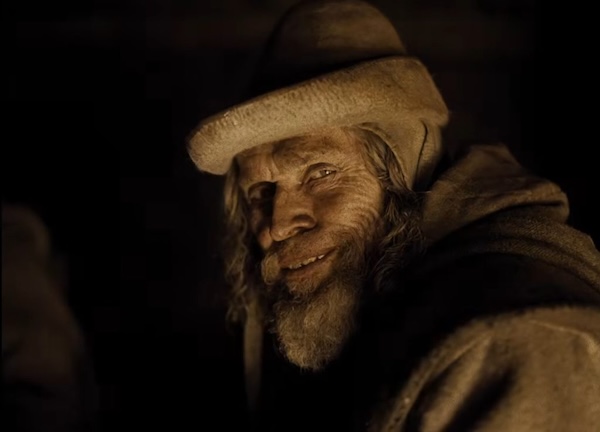 A dimly lit close-up of a grinning old man with a long grey beard and a pale felt hat, his deeply lined face picked out in warm light against a dark background.